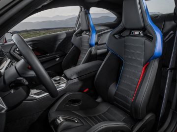 Binnenaanzicht van een BMW M2 met zwartleren sportstoelen met blauwe en rode accenten, middenconsole en dashboard. De auto heeft een modern en strak design.