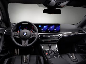 Binnenaanzicht van een BMW M2 met de nadruk op het stuur, het dashboard en de digitale displays. De cabine heeft een modern design met zwarte bekleding en koolstofvezelaccenten.