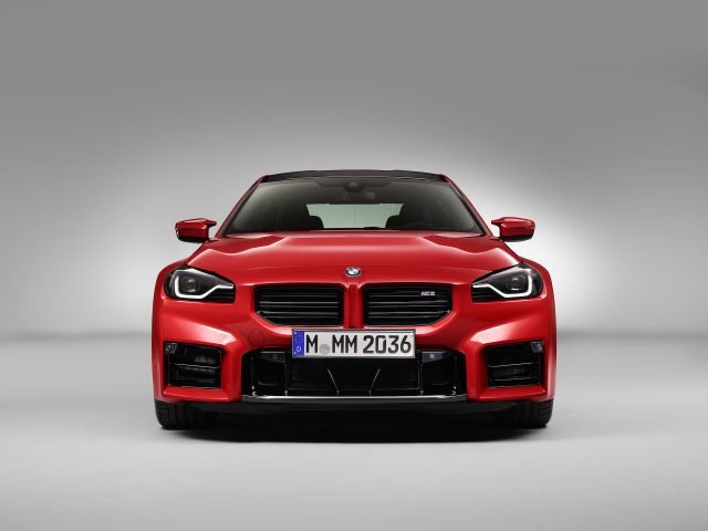 Vooraanzicht van een glanzend rode BMW M2 met een Duits kenteken "M MM 2036", tentoongesteld tegen een grijze achtergrond.