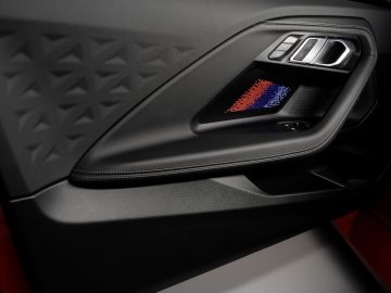 Close-up van het binnenpaneel van een BMW M2-autodeur met een geometrisch patroon, een bedieningscluster inclusief knoppen voor raam- en spiegelverstelling, en een decoratief element met rode, blauwe en zwarte strepen.