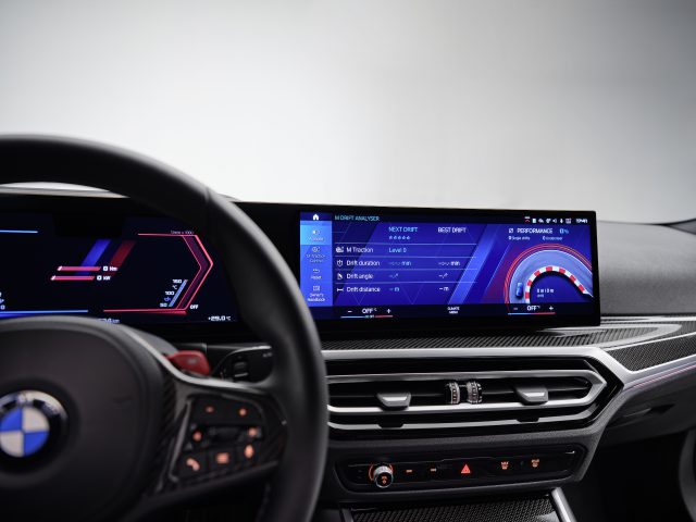 Binnenaanzicht van een BMW M2-autodashboard met een digitaal display met verschillende rijopties en prestatiegegevens. Het stuur met bedieningselementen is links gedeeltelijk zichtbaar.