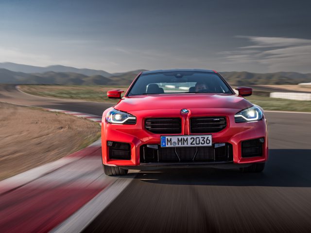 Een rode BMW M2-sportwagen snelt over het circuit met bergen op de achtergrond, vastgelegd vanaf de voorkant met een verbluffende bewegingsonscherpte.