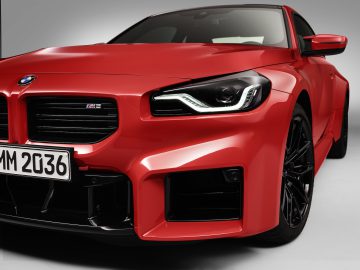 Close-up van een rode BMW M2-sportwagen, met de grille, de koplampen en een deel van de motorkap. De auto is voorzien van een kentekenplaat met "MM 2036" en zwarte details rond de grille.