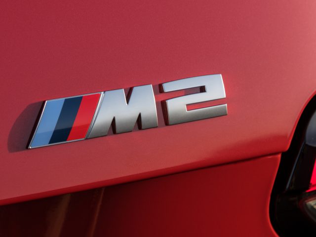 Close-up van het 'M2'-embleem van een rode auto, met de driekleurige strepen van de iconische BMW M-divisie naast de 'M' en het cijfer '2' in zilver, waardoor de auto duidelijk wordt gemarkeerd als een BMW M2.