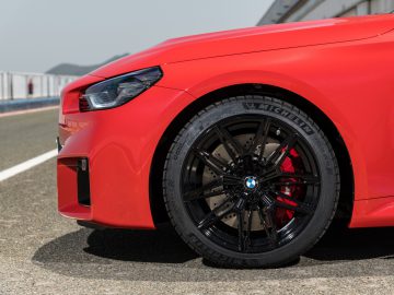 Close-up van het voorwiel van een rode BMW M2 op een weg. De zwarte lichtmetalen velg is voorzien van een Michelin-band en de auto is voorzien van een BMW-logo.