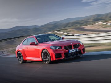 Een rode BMW M2-sportwagen rijdt op een racecircuit met bergen op de achtergrond onder een bewolkte hemel.