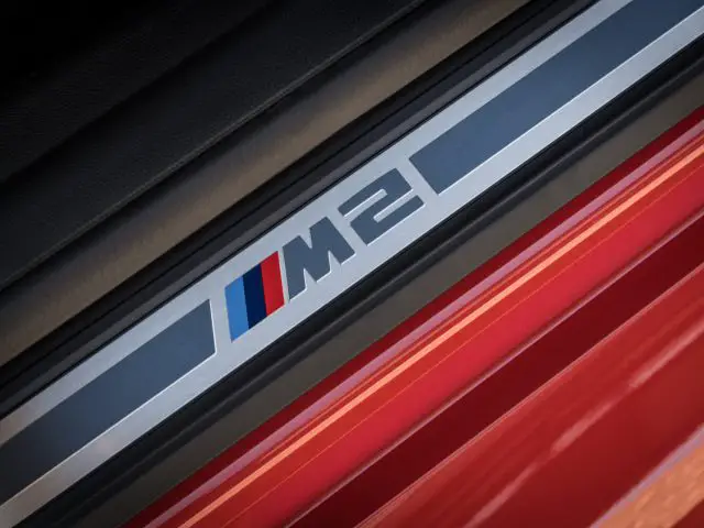 Close-upafbeelding van een BMW M2-instaplijst met het "M2"-logo in metallic met drie gekleurde strepen (blauw, paars en rood), geplaatst naast een rode carrosserie.