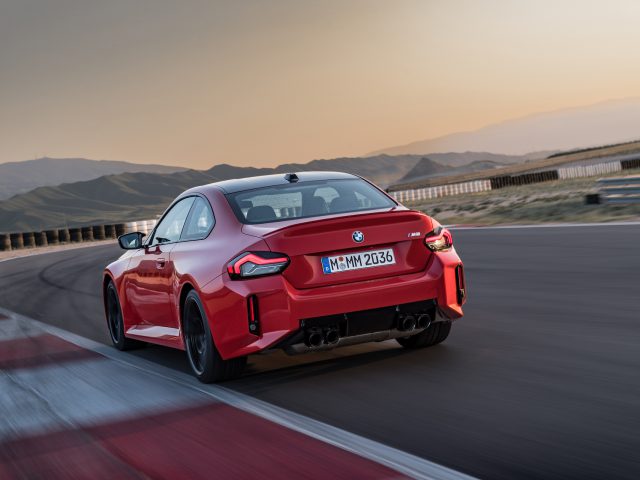 Een rode BMW M2-sportwagen rijdt op een racecircuit met bergen op de achtergrond tijdens een prachtige zonsondergang.