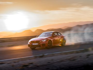 Een rode BMW M2-auto drijft bij zonsondergang over een racecircuit en creëert een rookwolk uit zijn banden. De achtergrond toont bergen en een wazige lucht.