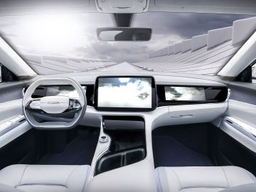 Futuristisch auto-interieur met twee stoelen, een groot centraal touchscreen en een strak dashboardontwerp. De voorruit toont een weergave van een moderne, gebogen architectonische structuur onder een gedeeltelijk bewolkte hemel, wat de mogelijkheden van autonoom rijden benadrukt.