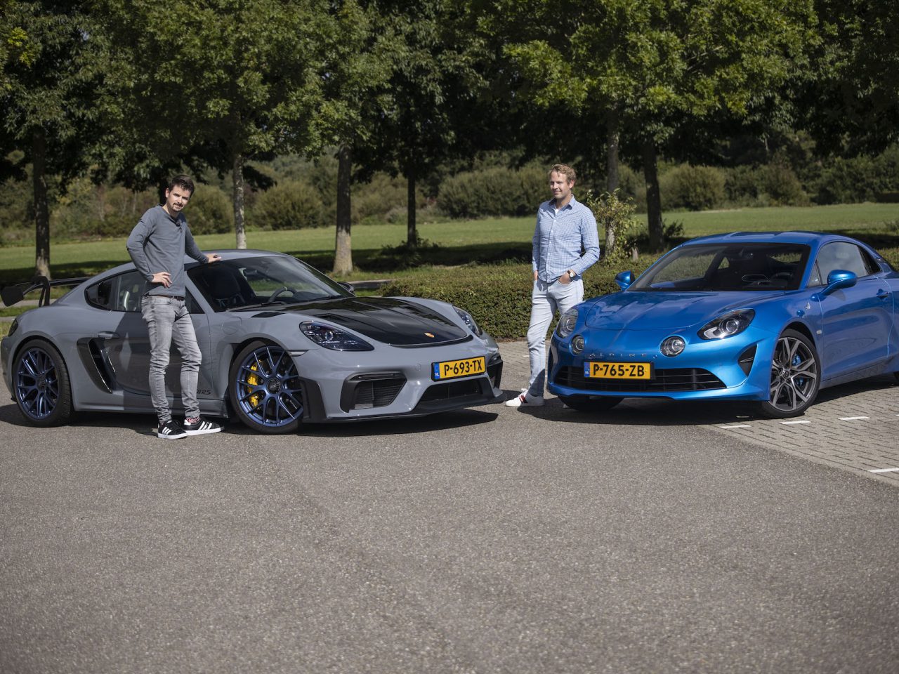 Twee mannen staan naast geparkeerde sportwagens op een buitenterrein; links een grijze compacte sportauto met middenmotor en rechts een blauwe auto. Op de achtergrond zijn bomen en grasvelden zichtbaar.