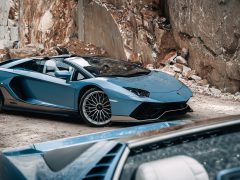 Een strakke blauwe Aventador met cabriokap staat geparkeerd op een rotsachtig industriegebied. Op de voorgrond is het interieur van een andere auto gedeeltelijk zichtbaar.