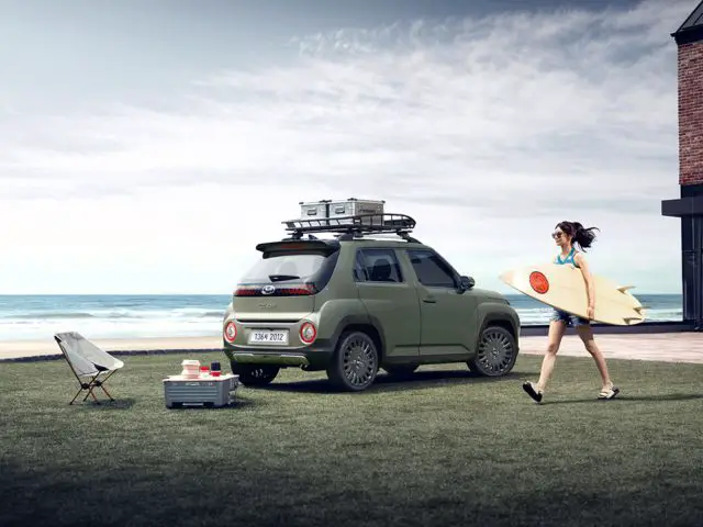 Een persoon met een surfplank loopt langs een groene Hyundai Casper die vlakbij een strand geparkeerd staat. Accessoires zijn op het dak van het voertuig gemonteerd en een opvouwbare stoel en koelbox staan vlakbij op het gras opgesteld.