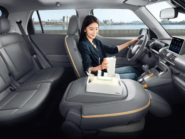 Een vrouw in een geparkeerde Hyundai Casper zit op de bestuurdersstoel, gebruikt een touchscreen terwijl ze een kopje vasthoudt. Het auto-interieur is modern met grijze stoelen en een consolevormig blad tussen de voorstoelen.