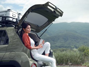 Een vrouw zit in de open kofferbak van een groene Hyundai Casper SUV, houdt een kopje vast en kijkt in de verte. Het voertuig staat buiten geparkeerd met groene heuvels en een bewolkte lucht op de achtergrond.