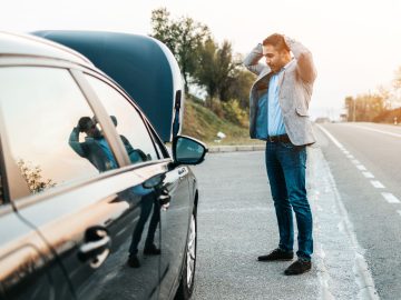 Een man staat met de handen op zijn hoofd langs de kant van de weg en kijkt met open motorkap naar zijn auto, die duidelijk spijt heeft dat hij verkeerde brandstof in de tank heeft gedaan.
