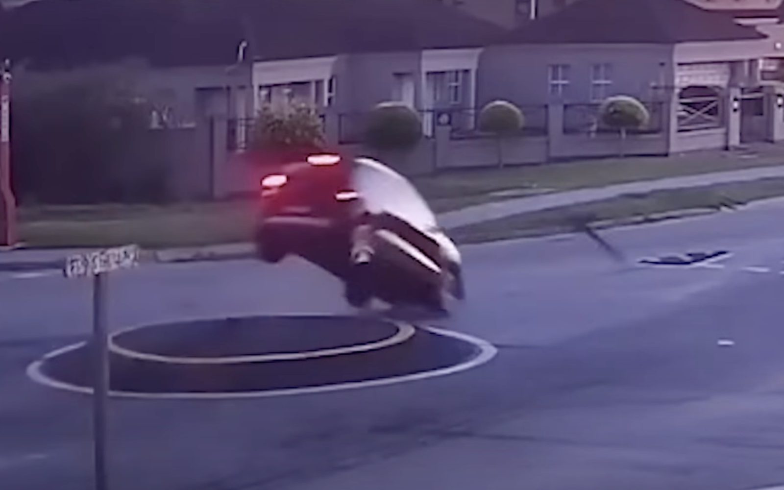 Een rode auto, bestuurd door idioten in de auto, wordt in de lucht gezien nadat hij over een verhoogd cirkelvormig gedeelte midden op een kruispunt in een buitenwijk is gereden. Op de achtergrond zijn nabijgelegen huizen en een wegwijzer zichtbaar.