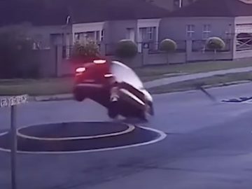 Een rode auto, bestuurd door idioten in de auto, wordt in de lucht gezien nadat hij over een verhoogd cirkelvormig gedeelte midden op een kruispunt in een buitenwijk is gereden. Op de achtergrond zijn nabijgelegen huizen en een wegwijzer zichtbaar.