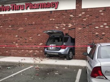Een auto botste tegen de bakstenen muur van een 'Drive-Thru Pharmacy', met de achterkant in het gebouw en stenen verspreid over de crashlocatie. Vlakbij staat een zilveren auto geparkeerd, wat bijdraagt aan het tafereel veroorzaakt door die idioten in de auto.