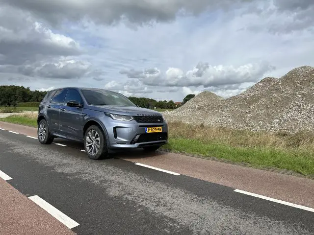 Een grijze Land Rover Discovery Sport P300e staat geparkeerd op een tweebaansweg met een stapel stenen en een bewolkte lucht op de achtergrond. Het voertuig heeft kenteken "P-029-PH".