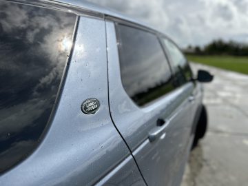 Close-up van een blauwe Land Rover Discovery Sport P300e met regendruppels op het raam en een bewolkte lucht weerspiegeld in het glas. Op de zijkant is het Land Rover-logo zichtbaar.