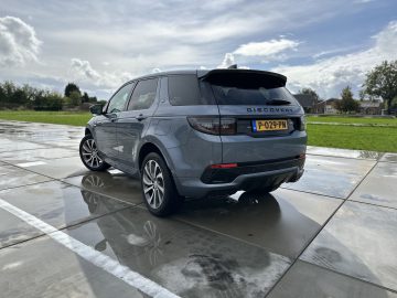 Een grijze Land Rover Discovery Sport P300e staat geparkeerd op een nat wegdek onder een bewolkte hemel.