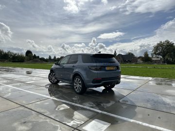 Een grijze Land Rover Discovery Sport P300e geparkeerd op een nat betonnen oppervlak en reflecteert het licht van een gedeeltelijk bewolkte lucht. Groen gras en huizen zijn zichtbaar op de achtergrond.