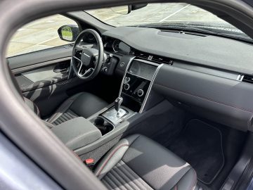 Interieur van een moderne Land Rover Discovery Sport P300e met zwart lederen stoelen, een centraal touchscreen-display en een versnellingspook in de middenconsole. Het stuur en het dashboard zijn ook zichtbaar.