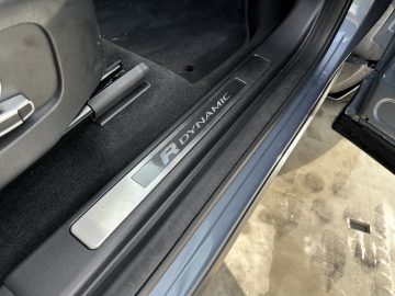 Auto-interieurdetail met een metalen deurdrempel met "R DYNAMIC" gegraveerd, zwart tapijt en een deel van de bestuurdersstoel in de Land Rover Discovery Sport P300e. De deur van het voertuig staat open, waardoor de natte grond buiten zichtbaar wordt.