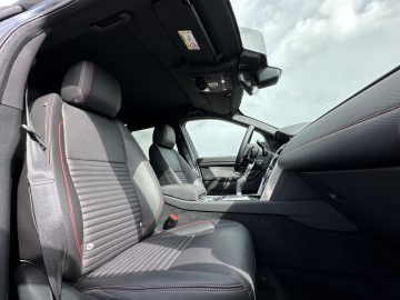 Binnenaanzicht van een Land Rover Discovery Sport P300e met de voorstoelen, het dashboard en de open passagiersdeur. De stoelen zijn zwart met rode stiksels en door de open deur en ramen is de lucht zichtbaar.