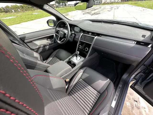 Interieur van een moderne Land Rover Discovery Sport P300e met de bestuurdersstoel, het stuur en het dashboard met touchscreen. De stoelen zijn voorzien van zwarte bekleding met rode stiksels. Het uitzicht is vanaf de achterbank.