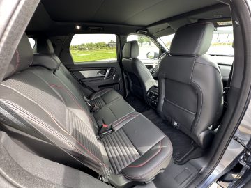 Binnenaanzicht van een Land Rover Discovery Sport P300e met de achterbank met zwart lederen bekleding en rode stiksels, de voorstoelen en een deel van de buitenkant van de auto door de open achterdeur op een zonnige dag.