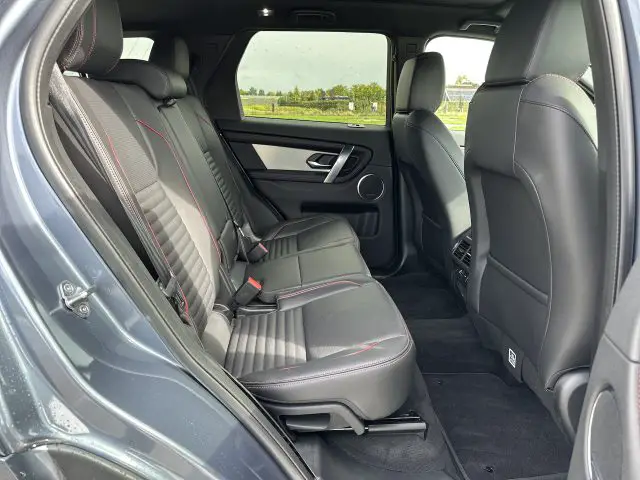 Een zicht op het achterste interieur van een Land Rover Discovery Sport P300e, met zwartleren stoelen met rode stiksels, deurpanelen en beenruimte op de achterbank. De foto is genomen vanaf de open passagiersdeur achteraan.