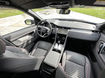 Uitzicht op het interieur van een auto vanaf de achterbank, met een modern dashboard met touchscreen, lederen stoelen en een stuur. Daglicht verlicht het strakke, strakke ontwerp van de Land Rover Discovery Sport P300e.