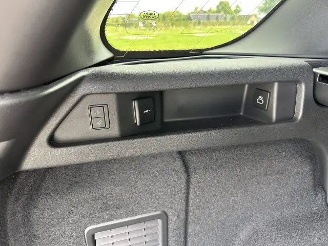 Close-up van de kofferbak van de Land Rover Discovery Sport P300e met bedieningsknoppen voor verschillende functies, een klein opbergvak en een deel van de achterruit met een schilderachtig uitzicht naar buiten.