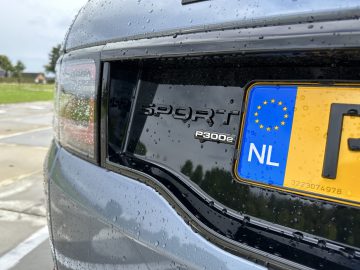 Close-up van de achterkant van een Land Rover Discovery Sport P300e met 'Sport'- en'P300e'-badges naast een kentekenplaat van de Europese Unie uit Nederland. Regendruppels zijn zichtbaar op het oppervlak van de auto.