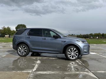 Een grijze Land Rover Discovery Sport P300e staat buiten op een nat oppervlak geparkeerd onder een bewolkte hemel, met wat groen zichtbaar op de achtergrond.