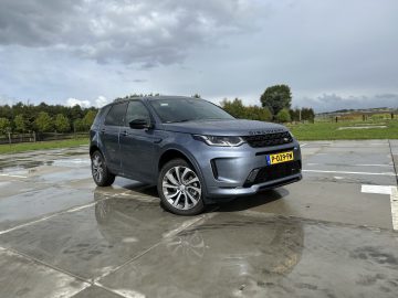 Een grijze Land Rover Discovery Sport P300e staat buiten geparkeerd op een nat betonnen oppervlak, met bomen en een bewolkte lucht op de achtergrond.