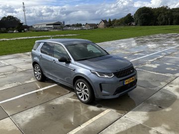 Een zilverkleurige Land Rover Discovery Sport P300e staat geparkeerd op een leeg parkeerterrein met een grasveld en gebouwen op de achtergrond. De grond is nat, wat wijst op recente regen.