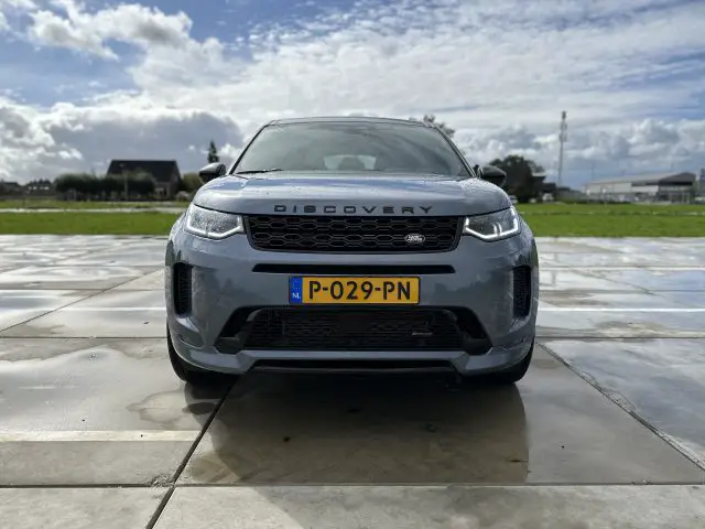 Een vooraanzicht van een grijs Land Rover Discovery Sport P300e-voertuig geparkeerd op een verhard oppervlak met een Europees kenteken "P-029-PN" onder een gedeeltelijk bewolkte hemel.