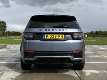 Een achteraanzicht van een grijze Land Rover Discovery Sport P300e SUV met Nederlands kenteken "P-029-PN", geparkeerd op een nat trottoir, met een grasachtig landschap op de achtergrond onder een bewolkte hemel.