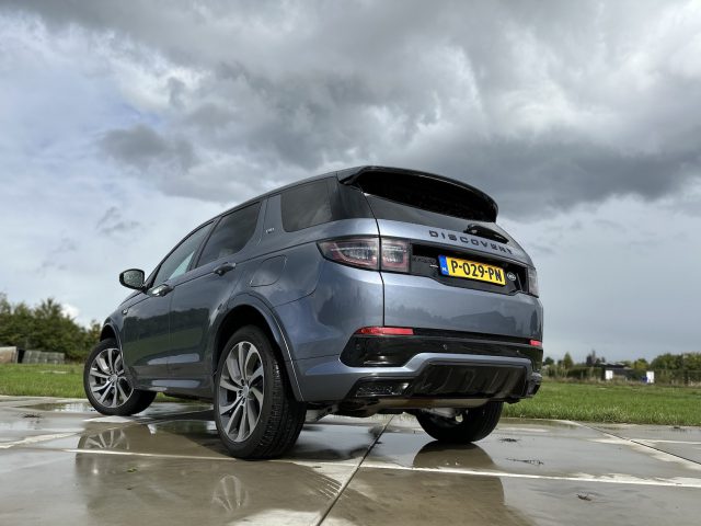 Een blauwe Land Rover Discovery Sport P300e SUV staat geparkeerd op een natte stoep met gras en een bewolkte lucht op de achtergrond. Het voertuig heeft een kenteken "P-029-PV".