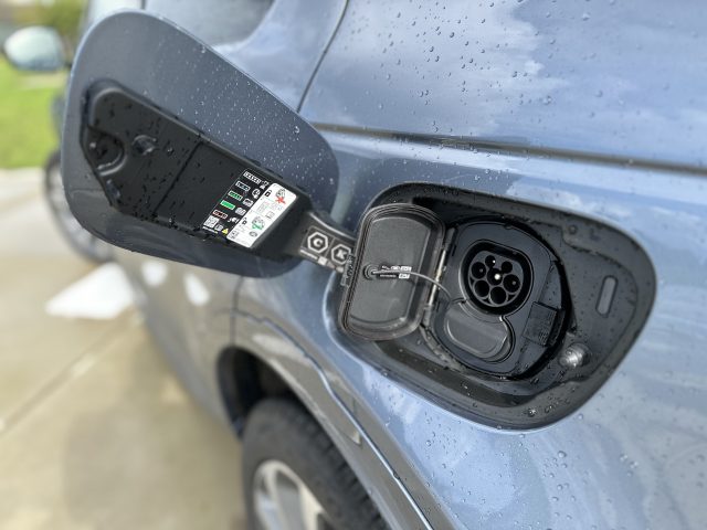 Close-up van een oplaadpoort voor elektrische voertuigen op een Land Rover Discovery Sport P300e, met de poortklep open en regendruppels op het oppervlak. De oplaadconnector is zichtbaar in de poort.