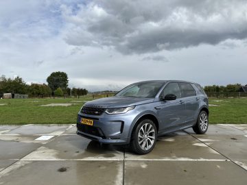 Een slanke zilveren Land Rover Discovery Sport P300e staat geparkeerd op een nat betonnen oppervlak, met een grasveld en een bewolkte lucht op de achtergrond.