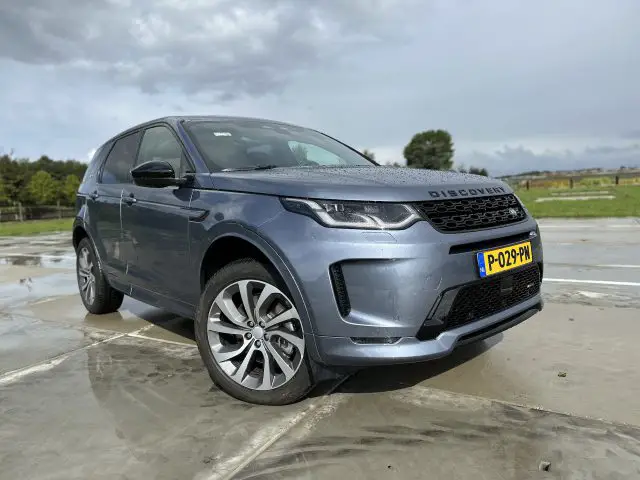 Een grijze Land Rover Discovery Sport P300e SUV staat geparkeerd op een nat betonnen oppervlak met een grasveld en een bewolkte lucht op de achtergrond.