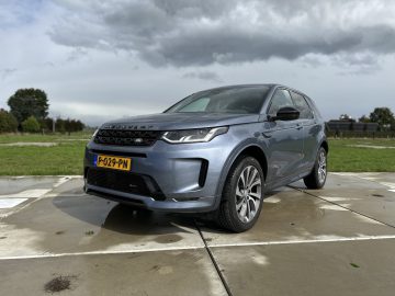 Een grijze Land Rover Discovery Sport P300e SUV geparkeerd op een betonnen ondergrond met een grasveld en bomen op de achtergrond onder een bewolkte hemel.
