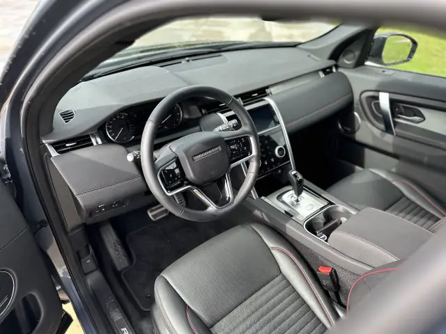 Binnenaanzicht van een Land Rover Discovery Sport P300e met de bestuurdersstoel, het stuur, het dashboard met digitale en analoge displays, en de middenconsole met bedieningselementen en versnellingspook.