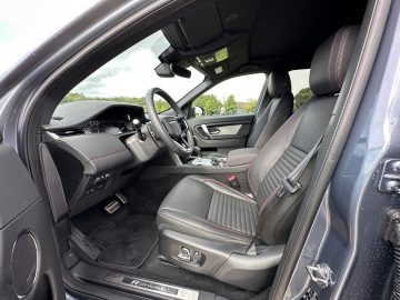 Binnenaanzicht van een Land Rover Discovery Sport P300e met de twee voorstoelen, het dashboard, het stuur en de middenconsole met versnellingspook. De stoelen zijn zwart met rood stiksel en het bestuurdersportier staat open.