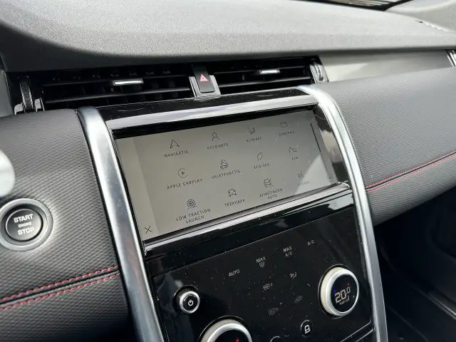 Close-up van een Land Rover Discovery Sport P300e-dashboard met een aanraakscherm met pictogrammen voor navigatie, ecogegevens, camera's en andere functies. Onder het scherm bevinden zich fysieke knoppen en draaiknoppen voor de klimaatregeling.