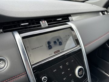 Close-up van een Land Rover Discovery Sport P300e-dashboard met een digitaal display met energie-informatie en verschillende bedieningsknoppen eronder. Het dashboard heeft een combinatie van gestructureerde en gladde oppervlakken.